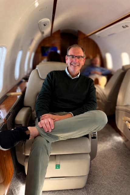 Smiling man in a green sweater sits cross-legged inside a luxury private jet cabin.